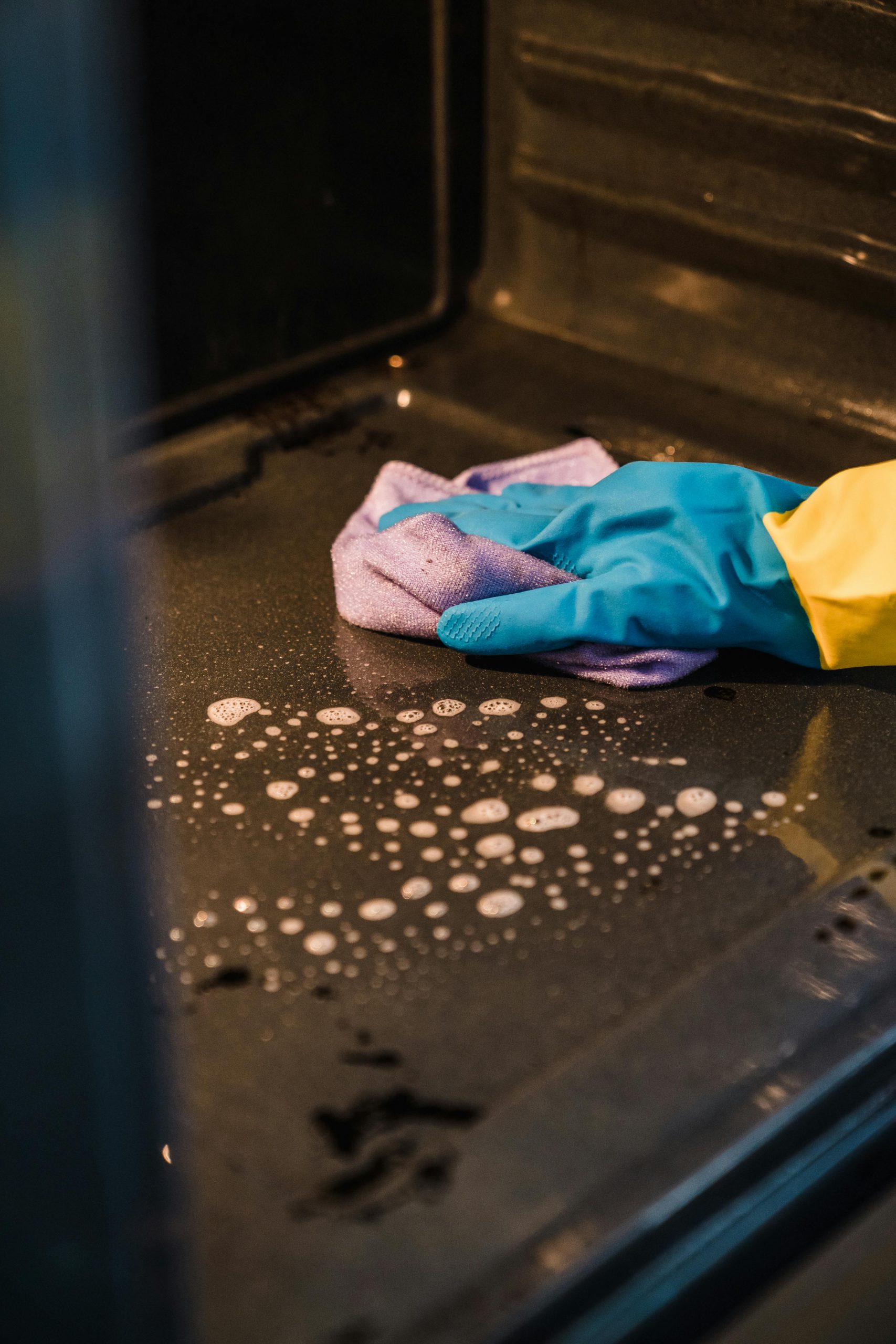 A gloved hand cleans an oven interior using a soapy cloth for a spotless finish.
